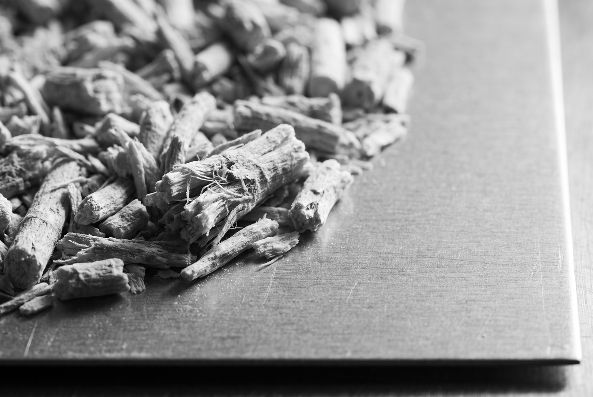 Fragments de racine d'ashwagandha séchée, fibreux et irréguliers, rassemblés en amas serré sur une tôle grise brossée